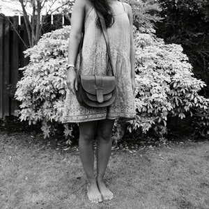 May include: A black and white image of a person wearing a patterned dress and a dark brown leather crossbody bag. The bag has a flap closure and a long strap. The person is standing barefoot on grass in front of a hedge.