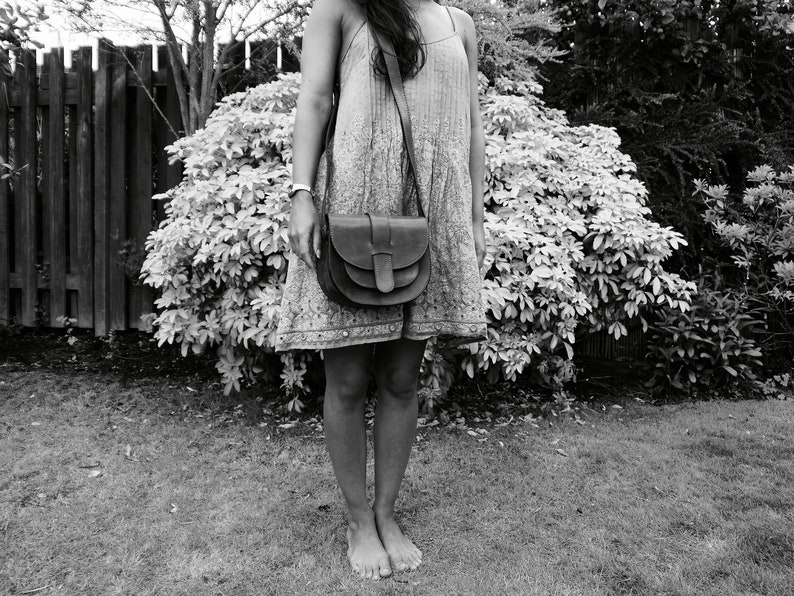 May include: A black and white image of a person wearing a patterned dress and a dark brown leather crossbody bag. The bag has a flap closure and a long strap. The person is standing barefoot on grass in front of a hedge.