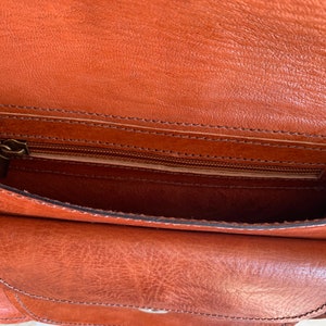 May include: Open view of a brown leather bag with a flap and a zippered pocket. The bag has a textured surface and visible stitching. The interior is a lighter shade of brown.