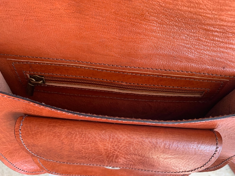 May include: Open view of a brown leather bag with a flap and a zippered pocket. The bag has a textured surface and visible stitching. The interior is a lighter shade of brown.