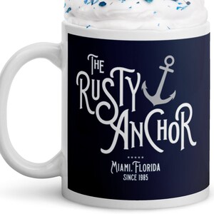 Puede incluir: Taza de cer&aacute;mica blanca con una banda azul marino con el texto "The Rusty Anchor" y un gr&aacute;fico de ancla. La taza est&aacute; llena de crema batida, chispas azules y una pajita a rayas rojas y blancas. Tambi&eacute;n es visible el texto "Miami, Florida Since 1985".