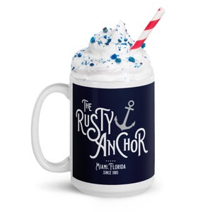 Puede incluir: Taza de cer&aacute;mica blanca con una banda azul marino con el texto "The Rusty Anchor" y un gr&aacute;fico de ancla. La taza est&aacute; llena de crema batida, chispas azules y una pajita a rayas rojas y blancas. Tambi&eacute;n es visible el texto "Miami, Florida Since 1985".