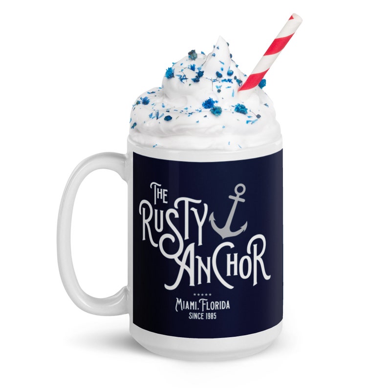 Puede incluir: Taza de cer&aacute;mica blanca con una banda azul marino con el texto "The Rusty Anchor" y un gr&aacute;fico de ancla. La taza est&aacute; llena de crema batida, chispas azules y una pajita a rayas rojas y blancas. Tambi&eacute;n es visible el texto "Miami, Florida Since 1985".