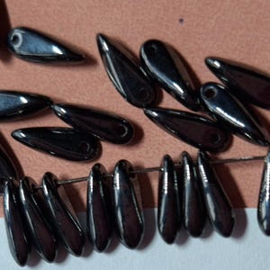 May include: A collection of dark, teardrop-shaped beads with a glossy finish. Some beads are strung on a thin wire, while others are scattered on a light brown surface. The beads have a small hole at the top for stringing.