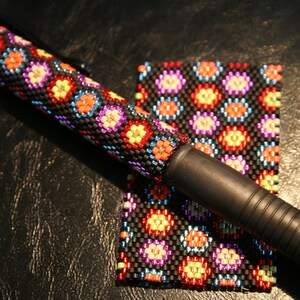 May include: A black pen with a beaded sleeve and matching square. The sleeve and square feature a repeating pattern of colorful floral designs in red, orange, yellow, blue, and purple. The pen has a black rubber grip.