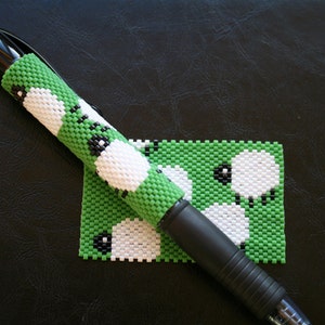 Op de afbeelding: Een zwarte pen met een groene en witte kralenhoes met een herhalend patroon van witte schapen met zwarte gezichten. De hoes is gemaakt van kleine kralen en is een leuke en unieke manier om een pen te personaliseren.