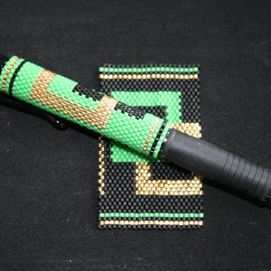 May include: A black and green beaded pen with gold accents. The pen is lying on a black surface next to a small black, green, and gold beaded square.
