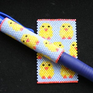 Peut inclure: Un stylo bleu avec un revêtement perlé présentant un motif répétitif de poussins jaunes sur un fond blanc et bleu. Le stylo est posé sur un petit carré perlé avec un motif similaire de poussins jaunes.