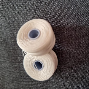 May include: Two spools of off-white thread are stacked on top of each other. The thread is tightly wound around a blue core. The background is a dark gray, textured fabric.