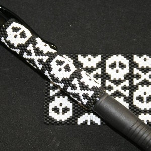 May include: A black pen with a white skull and crossbones pattern. The pen is lying on a black surface with a small square of fabric with the same pattern.