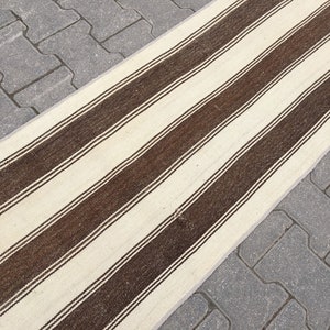 Puede incluir: Una alfombra larga de rayas blancas y marrones con un brillo sutil. La alfombra est&aacute; colocada sobre un camino de adoquines grises.