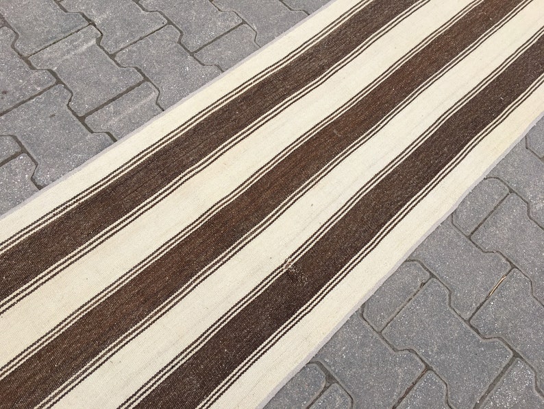 Puede incluir: Una alfombra larga de rayas blancas y marrones con un brillo sutil. La alfombra est&aacute; colocada sobre un camino de adoquines grises.