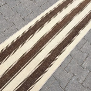 Puede incluir: Una alfombra larga y estrecha de rayas blancas y marrones. La alfombra est&aacute; hecha de un material tejido y tiene una textura sutil.