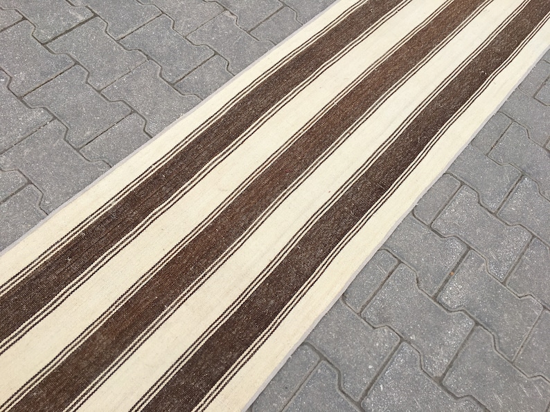Puede incluir: Una alfombra larga y estrecha de rayas blancas y marrones. La alfombra est&aacute; hecha de un material tejido y tiene una textura sutil.