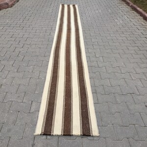 Puede incluir: Una alfombra de pasillo larga y estrecha con rayas marrones y blancas alternadas. La alfombra est&aacute; hecha de un material tejido y tiene un aspecto ligeramente desgastado.
