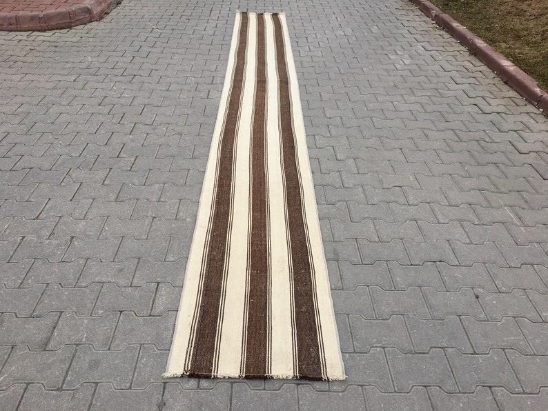 Puede incluir: Una alfombra de pasillo larga y estrecha con rayas marrones y blancas alternadas. La alfombra est&aacute; hecha de un material tejido y tiene un aspecto ligeramente desgastado.