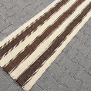 Puede incluir: Una alfombra larga de rayas blancas y marrones con un borde de flecos. La alfombra est&aacute; sobre una superficie de hormig&oacute;n gris.