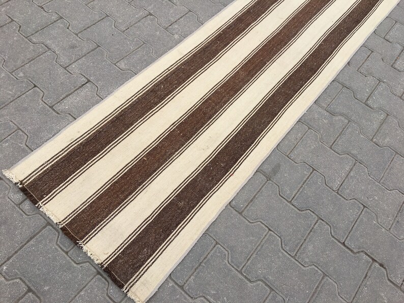 Puede incluir: Una alfombra larga de rayas blancas y marrones con un borde de flecos. La alfombra est&aacute; sobre una superficie de hormig&oacute;n gris.