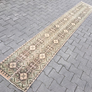 Puede incluir: Una alfombra larga y estrecha con un patrón geométrico en tonos marrones, verdes y beige. La alfombra está colocada sobre una superficie de hormigón gris.