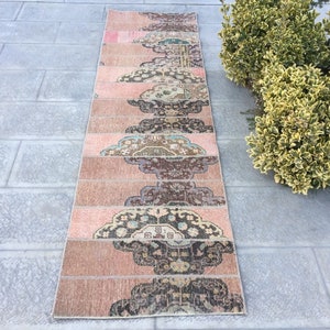 May include: A long, narrow rug with a pink background and a repeating pattern of brown, black, and blue floral designs. The rug is made of woven fabric and has a fringed edge.