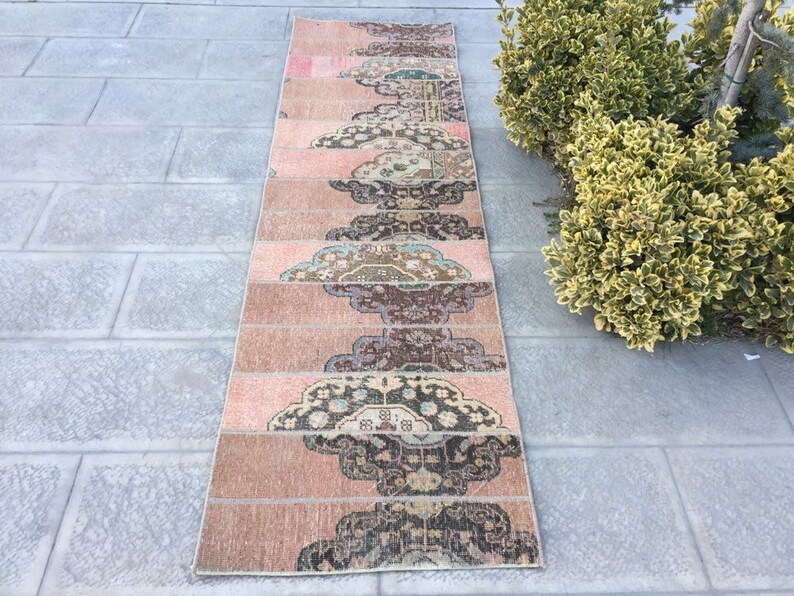 May include: A long, narrow rug with a pink background and a repeating pattern of brown, black, and blue floral designs. The rug is made of woven fabric and has a fringed edge.
