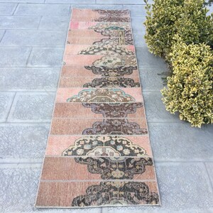 May include: A long, narrow rug with a pink background and a repeating pattern of dark brown, blue, and cream floral designs. The rug is made of woven fabric and has a fringed edge.