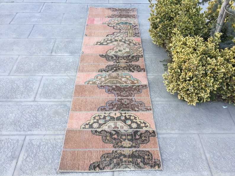 May include: A long, narrow rug with a pink background and a repeating pattern of dark brown, blue, and cream floral designs. The rug is made of woven fabric and has a fringed edge.