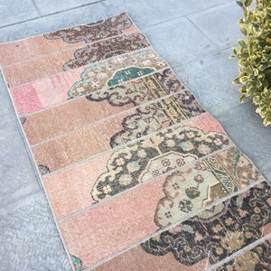 May include: A long, narrow rug with a pink and beige background. The rug is made of several strips of fabric sewn together, each with a different pattern. The patterns are mostly floral and geometric, in shades of brown, green, and black.
