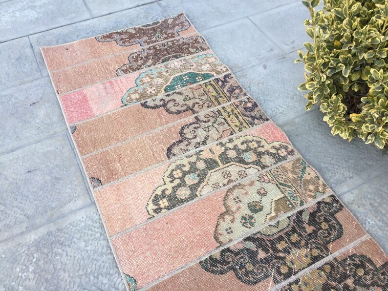 May include: A long, narrow rug with a pink and beige background. The rug is made of several strips of fabric sewn together, each with a different pattern. The patterns are mostly floral and geometric, in shades of brown, green, and black.