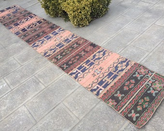 May include: A long, narrow rug with a colorful geometric pattern in shades of pink, blue, black, and green. The rug has a traditional, hand-woven look.