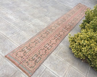 May include: A long, rectangular rug with a pink and green geometric pattern. The rug has a fringed edge and is lying on a gray tiled floor.