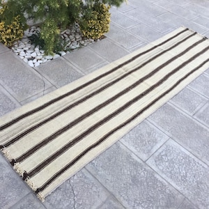 Puede incluir: Una alfombra larga y estrecha de rayas blancas y marrones con flecos en los extremos. La alfombra está sobre un suelo de baldosas grises.