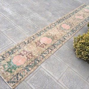 May include: A long, narrow rug with a beige background and a repeating pattern of pink, green, and brown floral motifs. The rug is lying on a gray tiled floor.