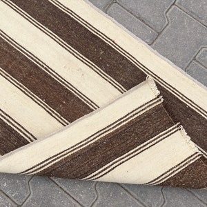 Puede incluir: Una alfombra de rayas blancas y marrones con borde de flecos. La alfombra est&aacute; doblada por la mitad y est&aacute; sobre una superficie de hormig&oacute;n gris.