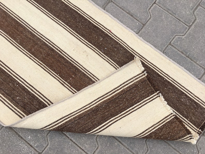 Puede incluir: Una alfombra de rayas blancas y marrones con borde de flecos. La alfombra est&aacute; doblada por la mitad y est&aacute; sobre una superficie de hormig&oacute;n gris.