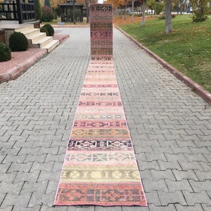 May include: A long, narrow, pink and beige runner rug with a traditional Turkish kilim pattern. The rug is laid out on a gray brick walkway.