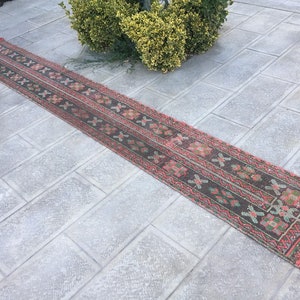 May include: A long, narrow runner rug with a traditional design. The rug features a dark brown central field with repeating floral and geometric patterns in red, pink, and green. The edges are trimmed in red.