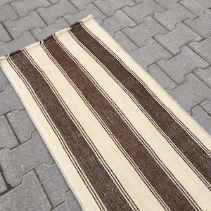 Puede incluir: Una alfombra larga y estrecha de color crema con rayas marrones. La alfombra est&aacute; sobre una superficie de hormig&oacute;n gris.