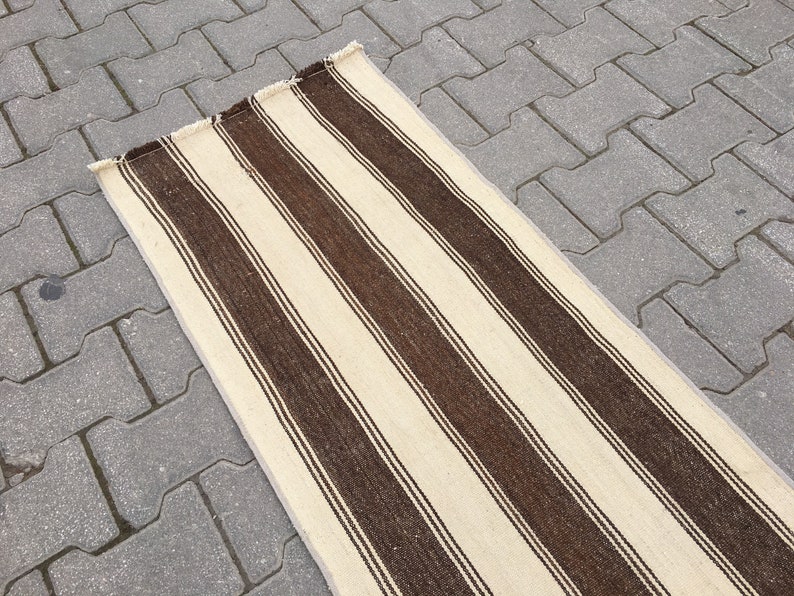 Puede incluir: Una alfombra larga y estrecha de color crema con rayas marrones. La alfombra est&aacute; sobre una superficie de hormig&oacute;n gris.
