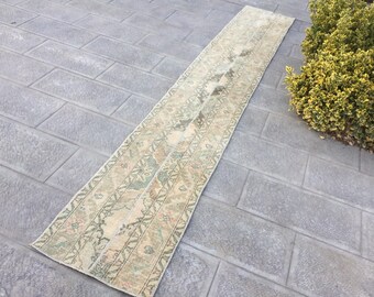 May include: A long, narrow, beige and green patterned rug with a floral design. The rug is laid out on a gray tiled floor.