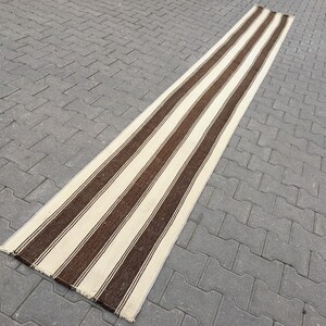 Puede incluir: Una alfombra larga y estrecha de rayas blancas y marrones con un borde de flecos. La alfombra est&aacute; sobre una superficie de hormig&oacute;n gris.