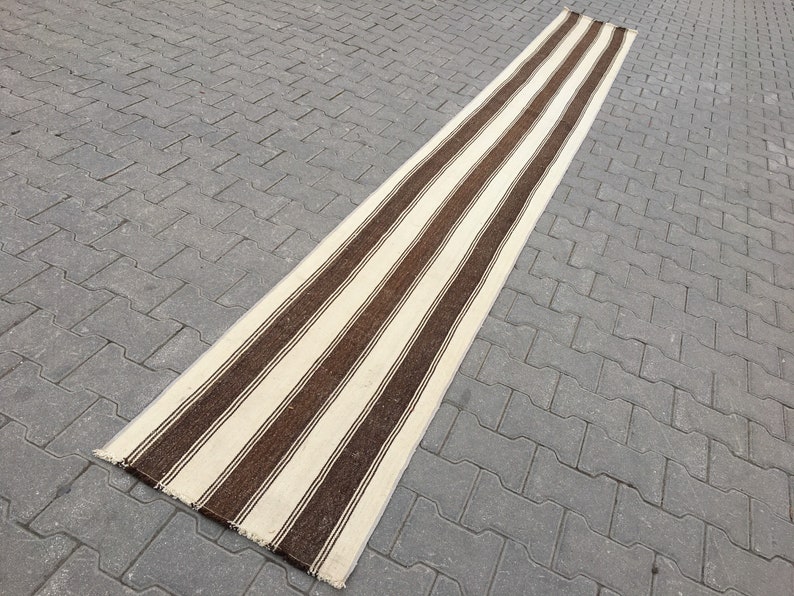 Puede incluir: Una alfombra larga y estrecha de rayas blancas y marrones con un borde de flecos. La alfombra est&aacute; sobre una superficie de hormig&oacute;n gris.