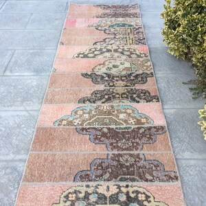 May include: A long, narrow rug made from multiple strips of fabric sewn together. The rug has a pink, brown, and cream color scheme with a floral pattern. The rug is laid out on a gray tiled floor.
