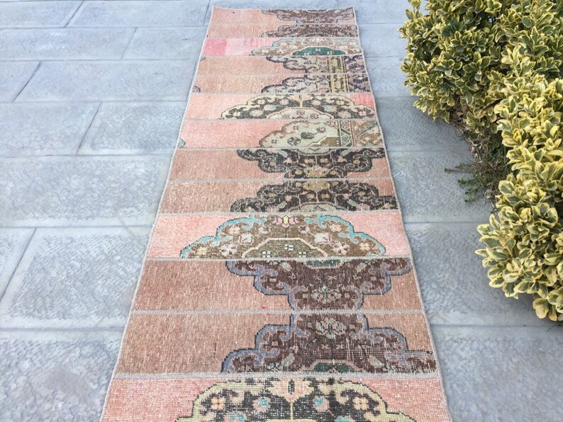 May include: A long, narrow rug made from multiple strips of fabric sewn together. The rug has a pink, brown, and cream color scheme with a floral pattern. The rug is laid out on a gray tiled floor.