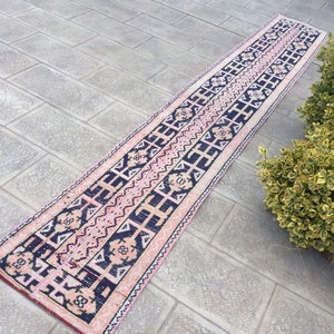 Puede incluir: Una alfombra larga de color rosa y negro con un diseño geométrico. La alfombra está colocada sobre un suelo de baldosas grises.