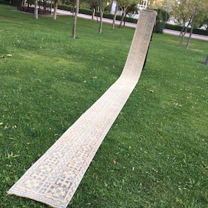 May include: A long, cream-colored rug with a geometric pattern in shades of blue, brown, and beige. The rug is laid out on a green grassy lawn.