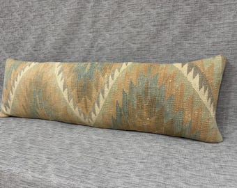 May include: Rectangular decorative pillow with a geometric pattern in shades of beige, brown, and teal. The pillow has a long, narrow shape and is placed on a gray textured surface. The pattern features repeating zigzag shapes.