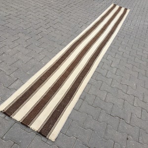 Puede incluir: Una alfombra larga y estrecha de rayas blancas y marrones con un borde de flecos. La alfombra est&aacute; sobre una superficie de hormig&oacute;n gris.