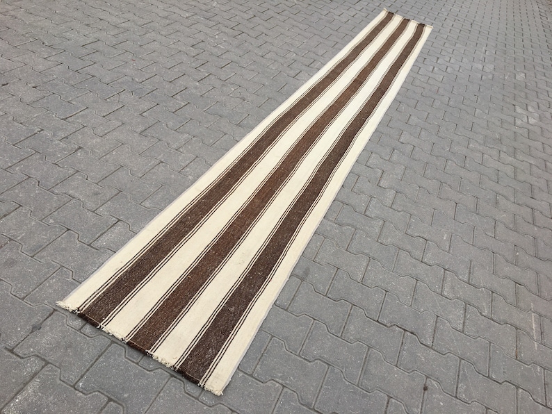 Puede incluir: Una alfombra larga y estrecha de rayas blancas y marrones con un borde de flecos. La alfombra est&aacute; sobre una superficie de hormig&oacute;n gris.
