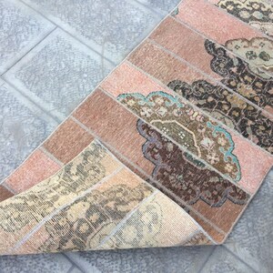 May include: A vintage runner rug with a pink and brown color scheme. The rug has a geometric pattern with a floral design. The rug is folded in half, showing the back side of the rug.